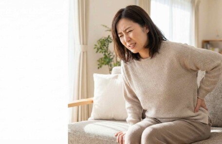 リビングのソファで股関節の痛みを感じて顔をしかめる40代の日本人女性